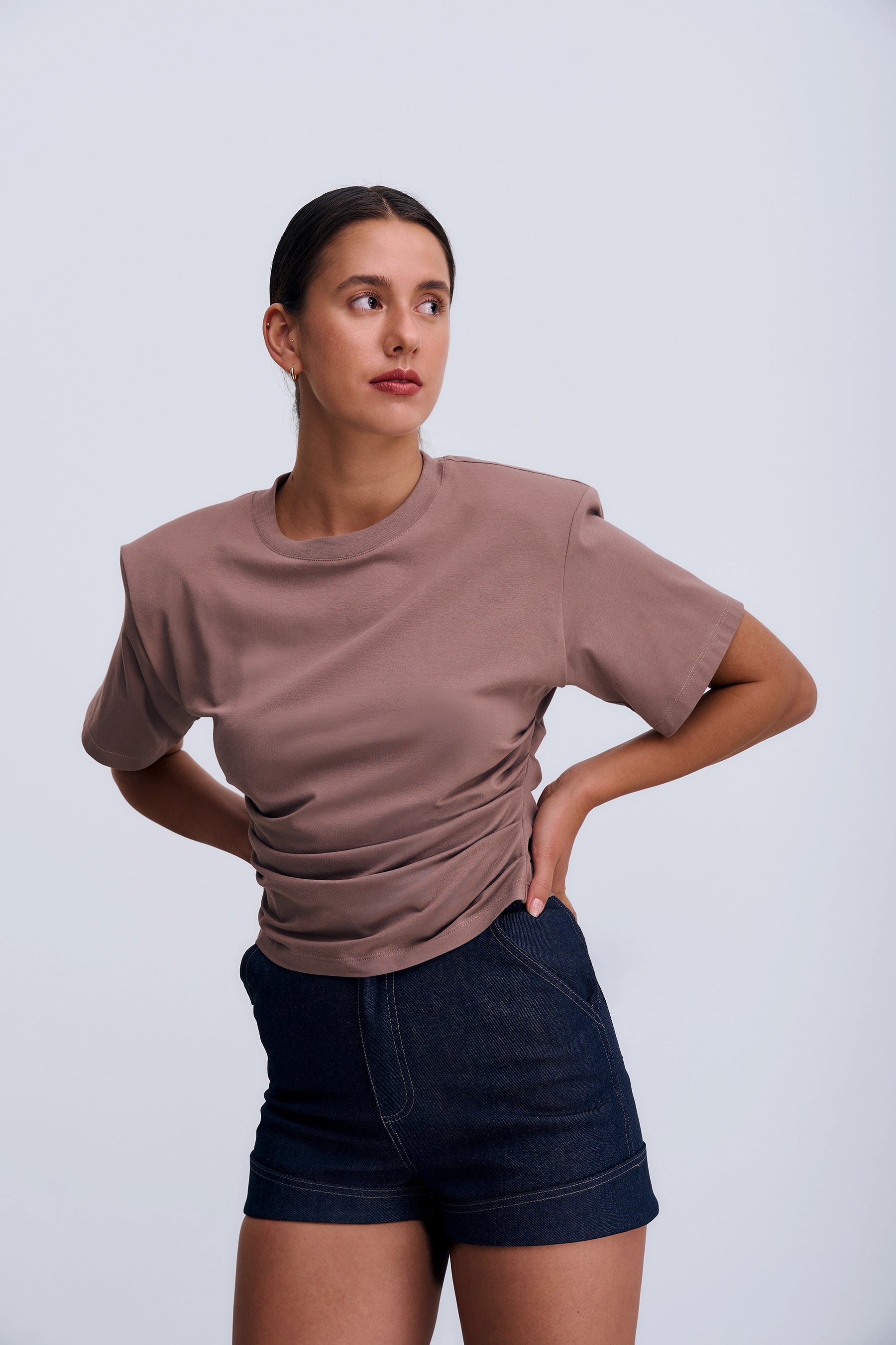 Modelka w Siena T‑shirt Taupe – minimalistyczny T‑shirt z poduszkami na ramionach, nowoczesny krótki fason z eleganckim akcentem przy ramionach.