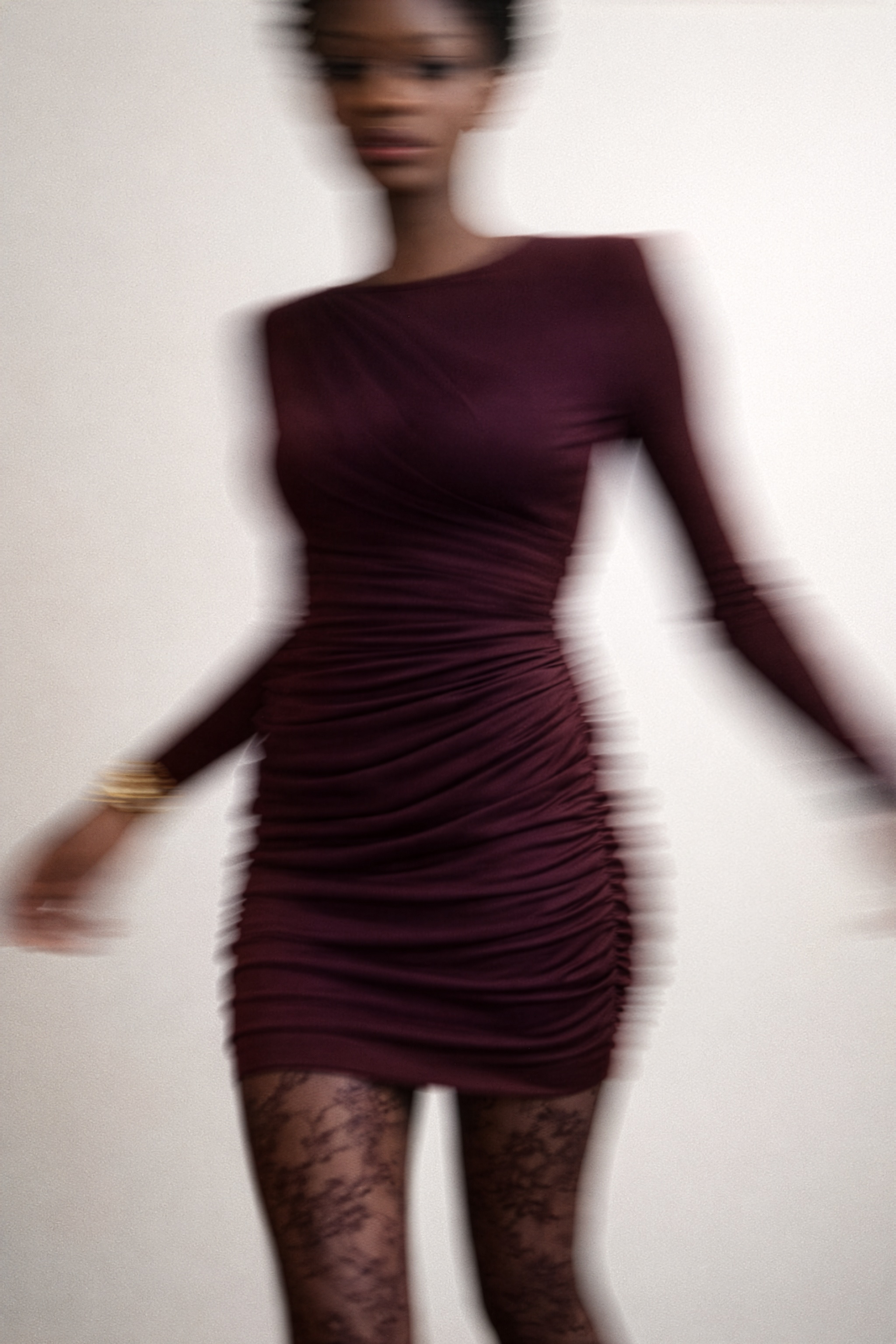 Modelka w bordowej, dopasowanej sukience z długim rękawem, w dynamicznym, lekko rozmytym ujęciu na jasnym tle.